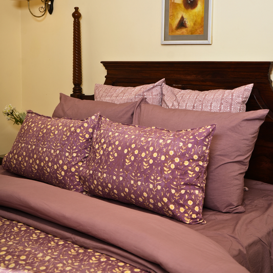 Bed with purple floral bedding and pillows against a beige wall.