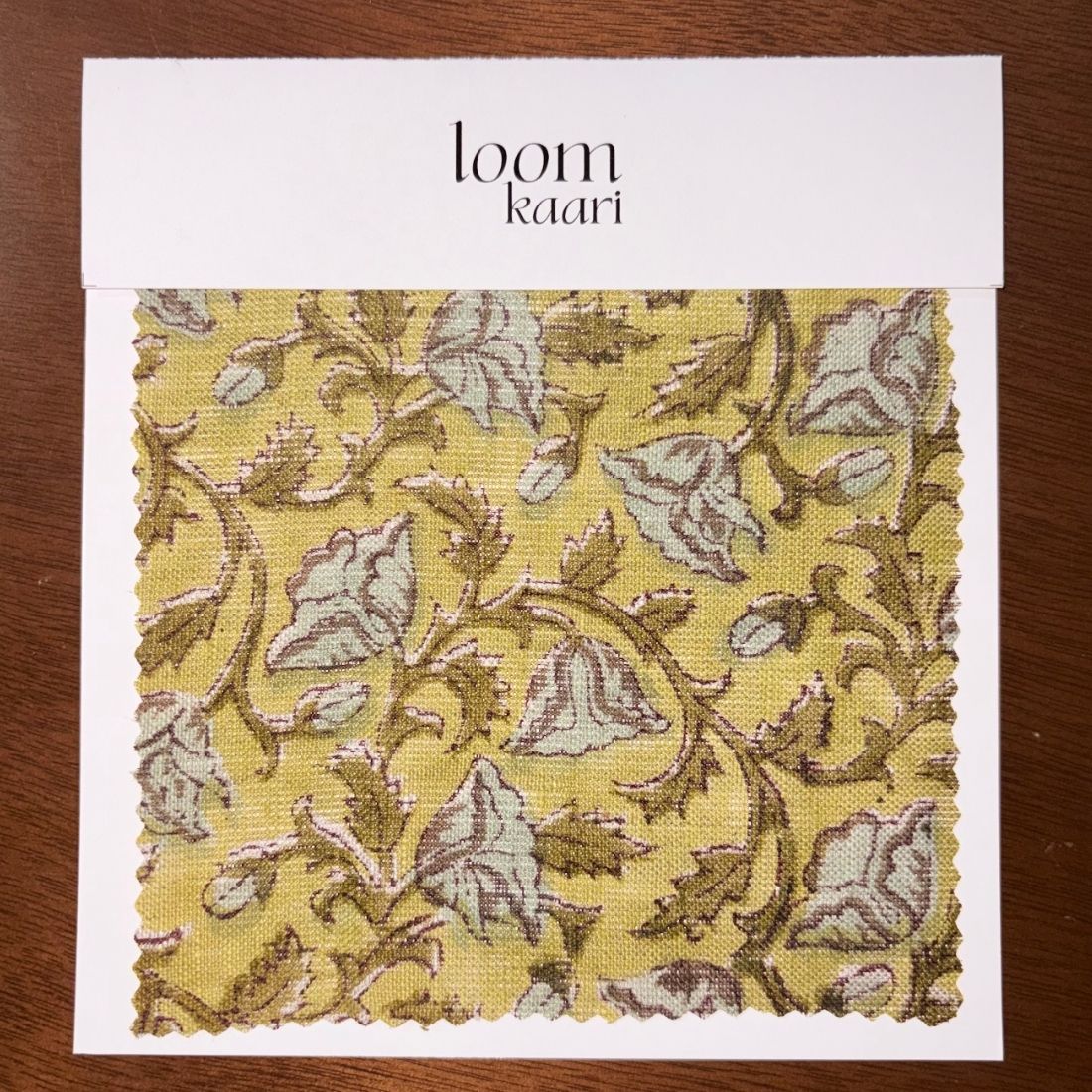 Fabric sample with leaf pattern on a white card labeled 'loom Kaari' on a wooden surface.