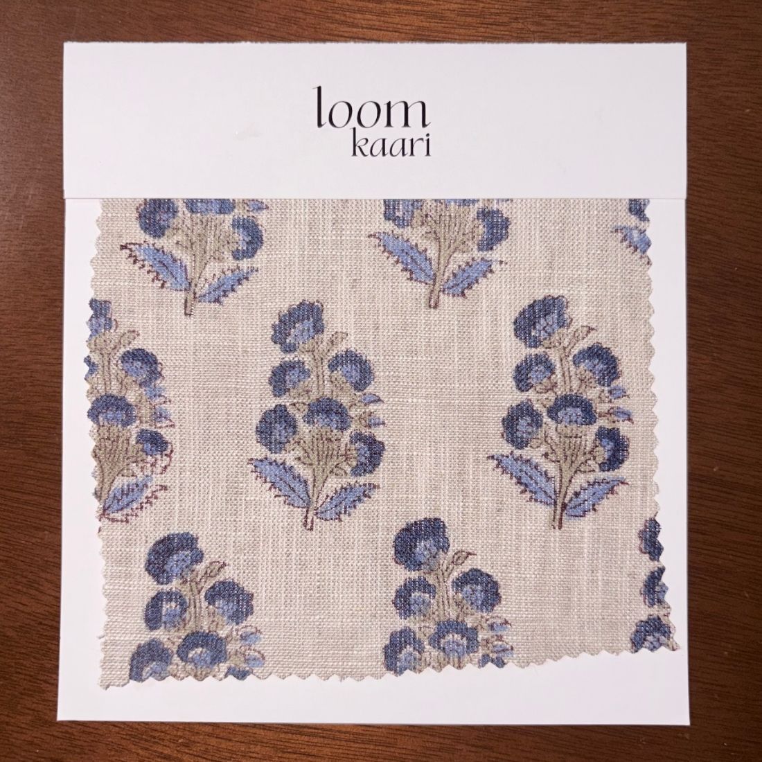 Fabric sample with blue floral pattern on a white card labeled 'loom kaari' on a wooden surface.