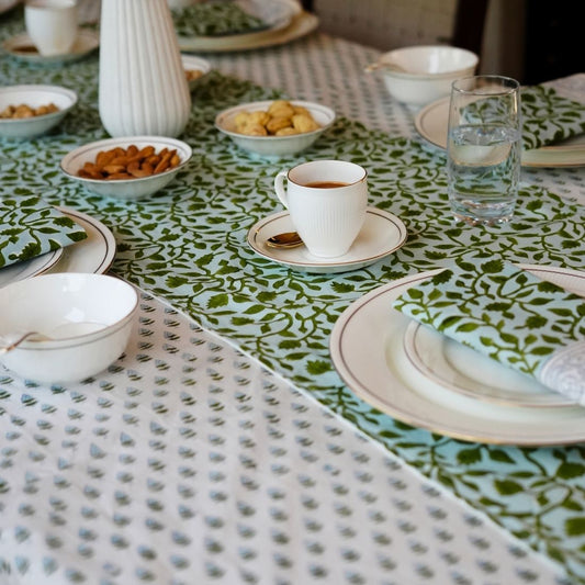 Table setting with a green and white patterned tablecloth, table runner, napkins, cups, saucers, and plates.