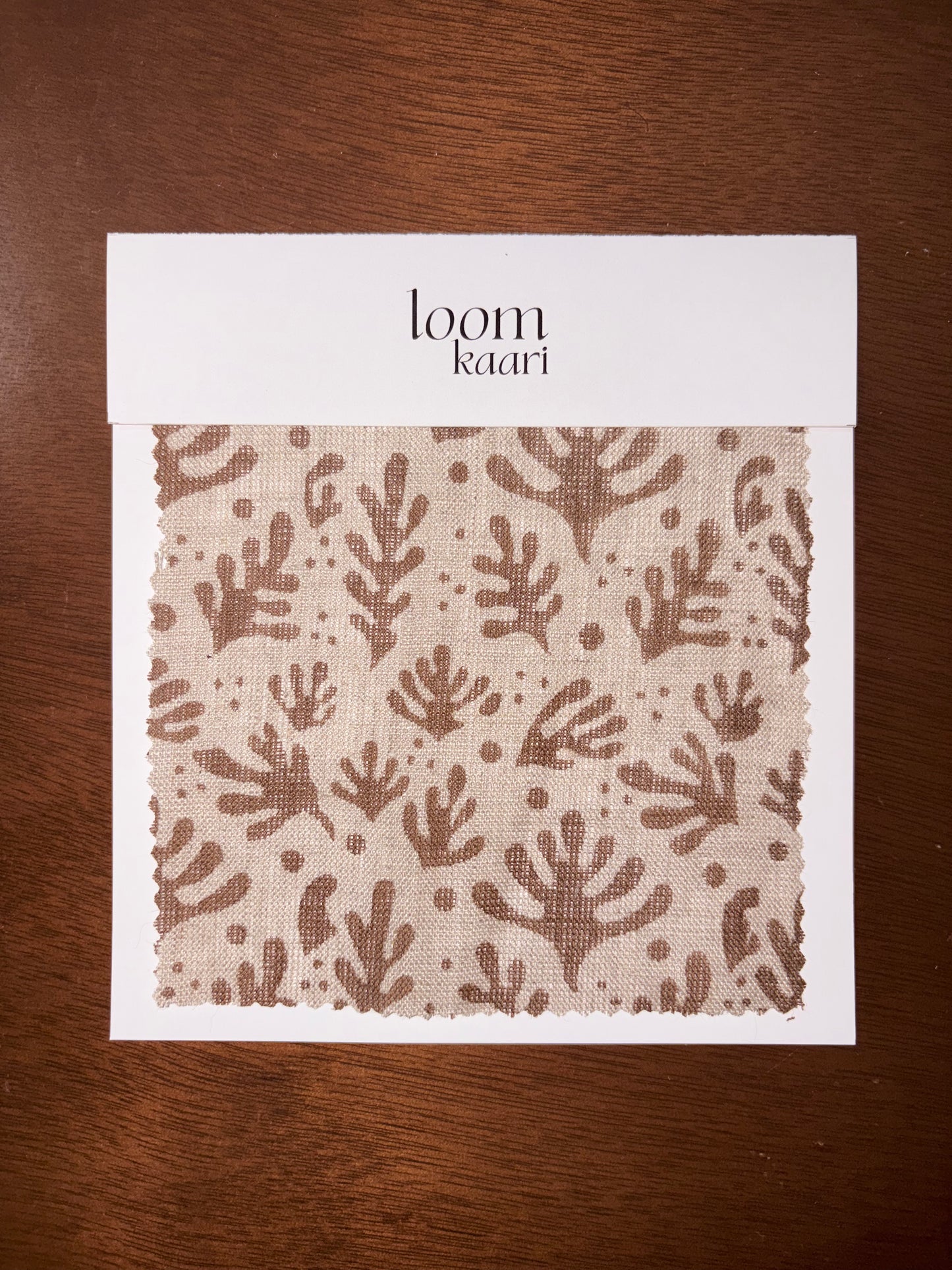 Fabric sample with a leaf pattern on a white card labeled 'loom Kaari' against a wooden background