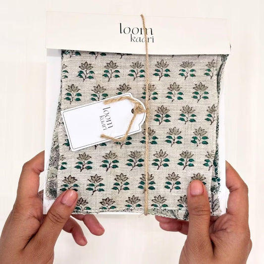 A person's hands hold a packaged fabric item wrapped in a white paper sleeve with the brand name 'loom kaari' printed at the top. The fabric has a beige base with a repeating green and brown floral block print, tied with a jute twine string and a small paper tag.