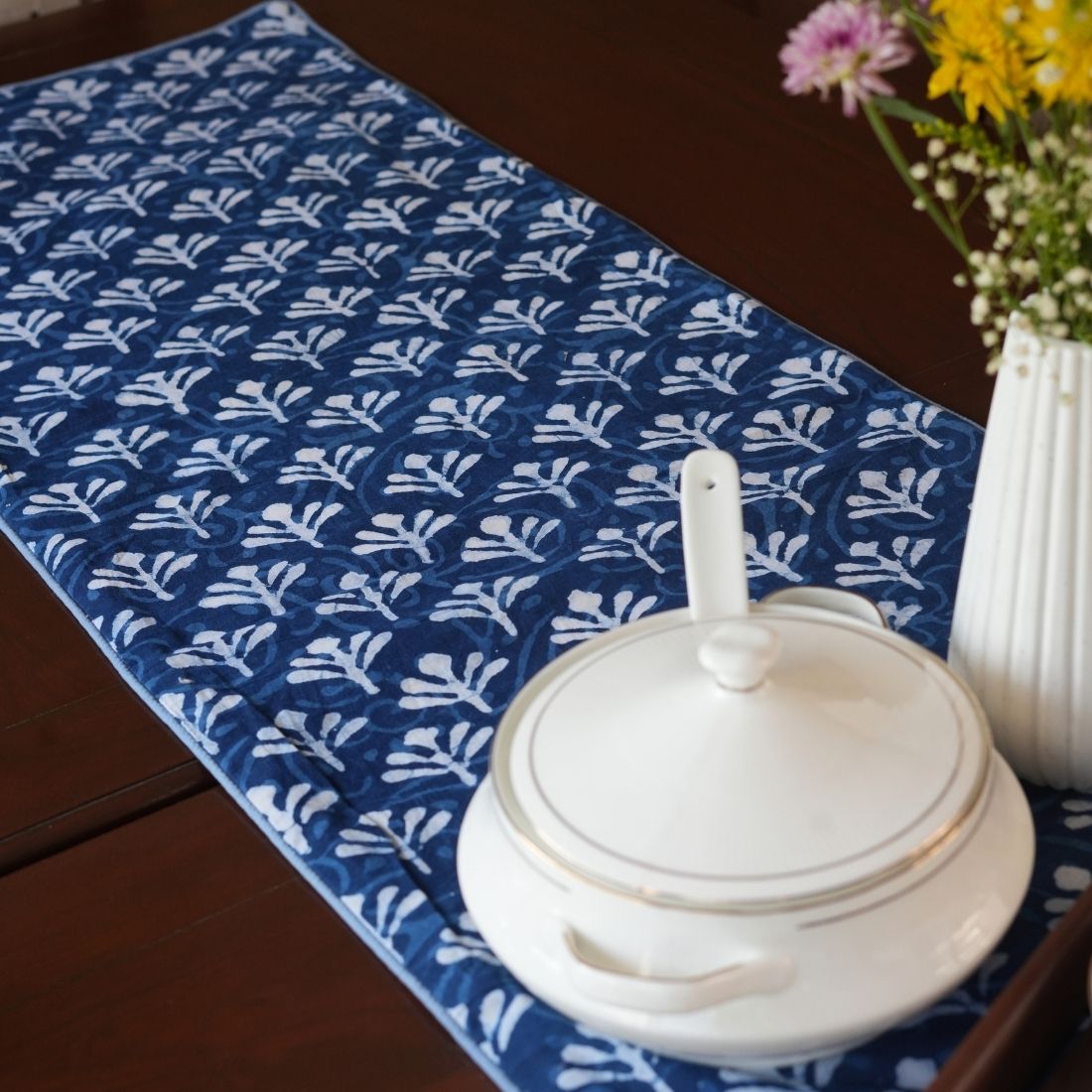 White ceramic pot on a blue patterned tablerunner with a vase of flowers in the background.