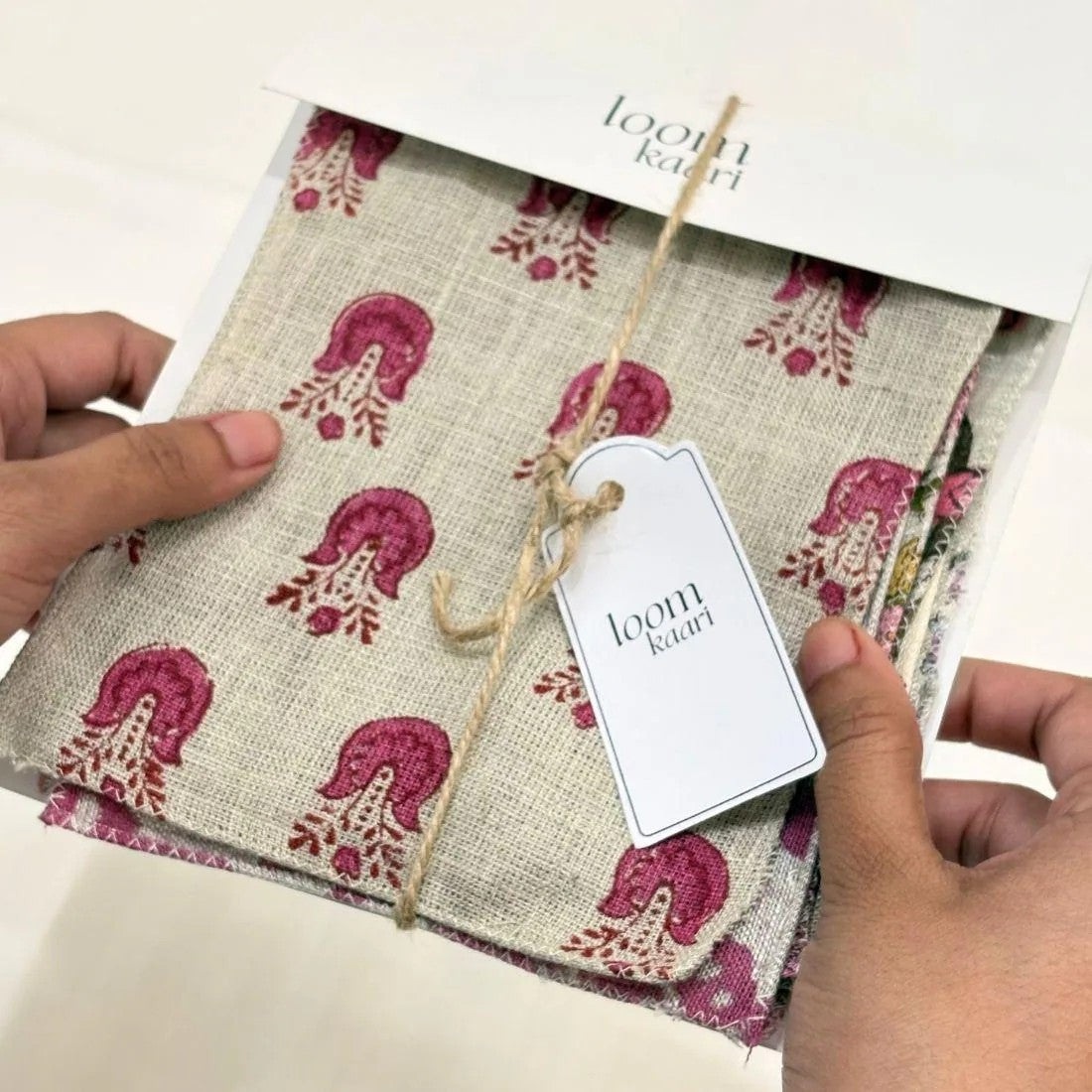 A person's hands holding a folded fabric bundle wrapped in a white paper sleeve and tied with a jute twine tag. The fabric has a natural beige linen-like base with a repeating magenta and cream floral pattern.