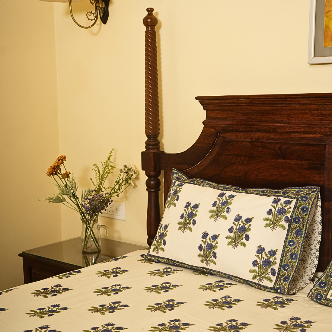 Bed with floral patterned bedding and pillow, nightstand with flowers, wooden headboard.