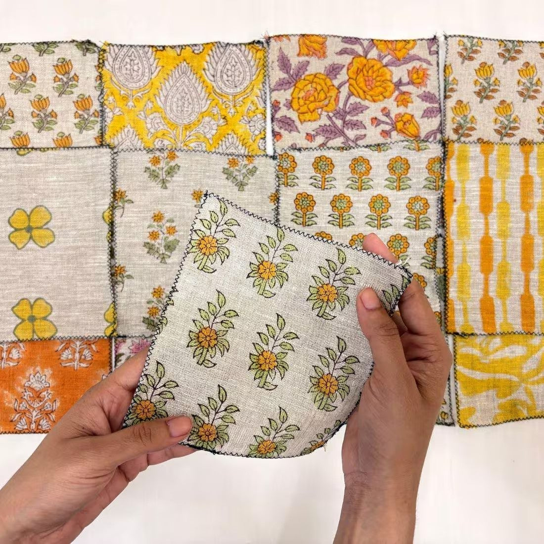 A person's hands hold a square fabric swatch featuring a yellow and green floral pattern on a light beige linen background, with a grid of other colorful patterned fabric swatches displayed in the background.