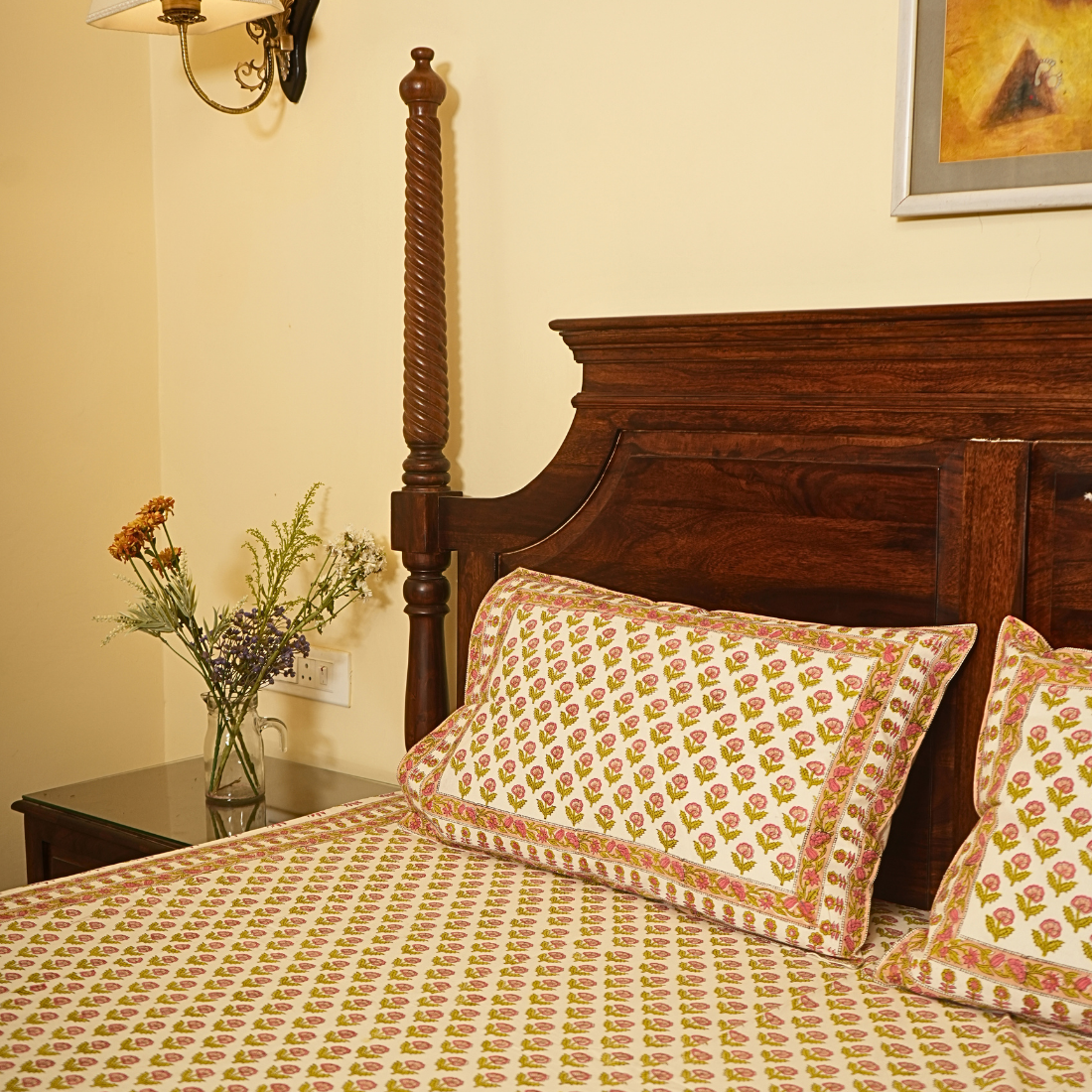 Bedroom with wooden bed and floral bedding, nightstand with flowers, and wall art.