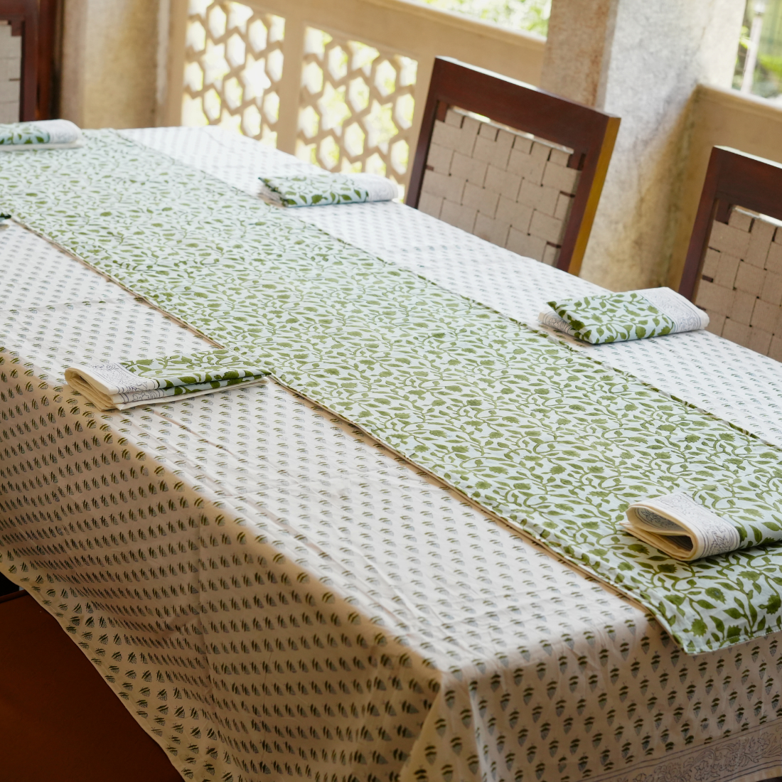 Table setting with green and beige tablecloth, napkins, and chairs in a bright room.