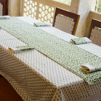 Table setting with green and beige tablecloth, napkins, and chairs in a bright room.