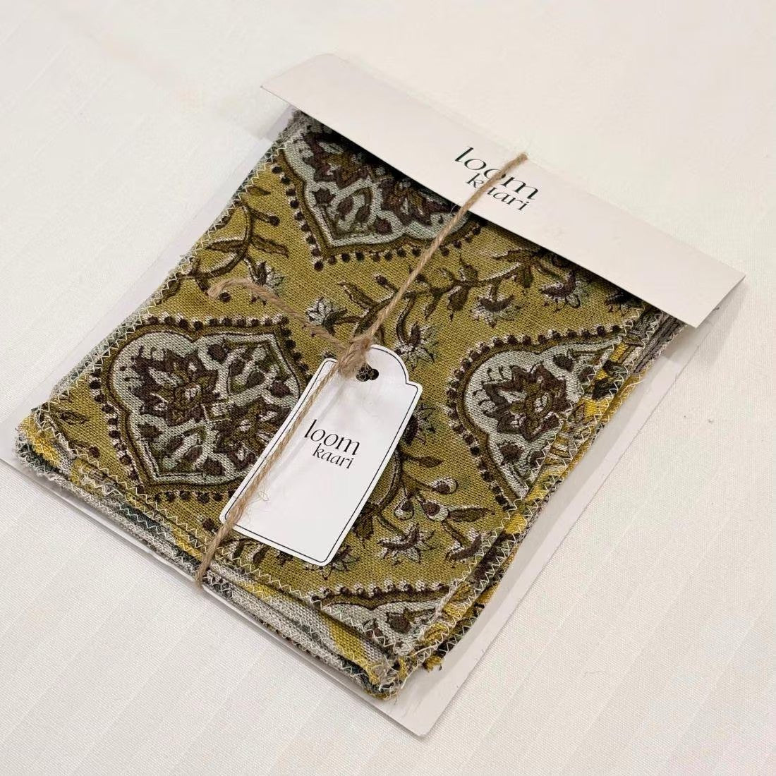 A packaged fabric or textile product with a printed white header card and hanging tag, photographed on a plain light background. The visible fabric has an ornate paisley-style pattern in olive green, beige, and brown tones.