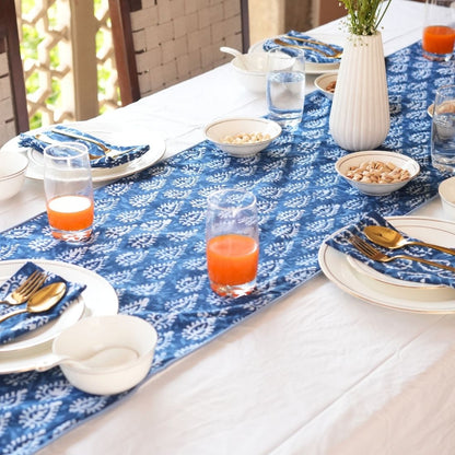 Dining table set with blue floral table runner, white plates, glasses of orange juice, and silverware.