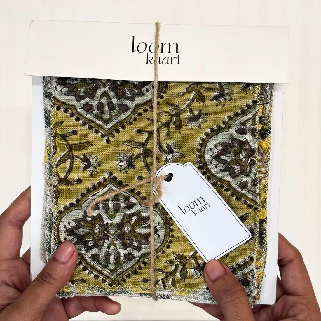 A person's hands holding a packaged fabric sample from 'loom khari', showing a mustard yellow and brown patterned textile with a white tag attached.