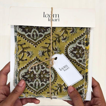 A person's hands holding a packaged fabric sample from 'loom khari', showing a mustard yellow and brown patterned textile with a white tag attached.