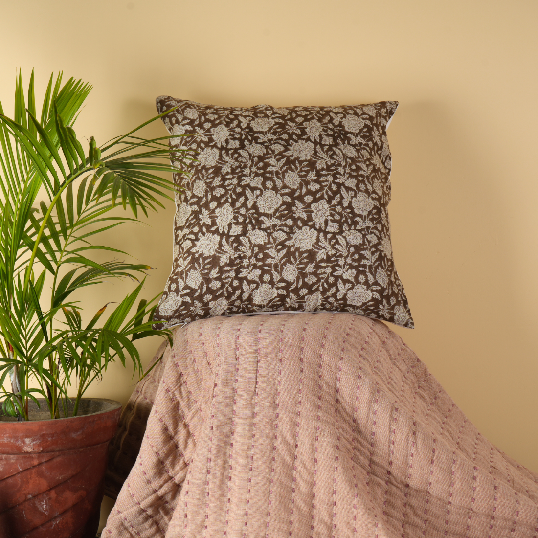 Decorative pillow with floral pattern on a bed next to a potted plant against a beige wall.