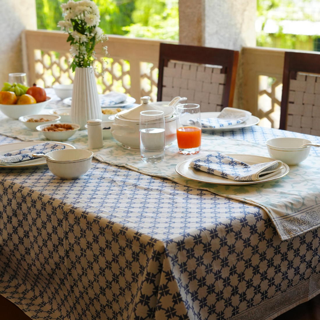 Dining table set with plates, glasses, and a vase on a patterned tablecloth outdoors.