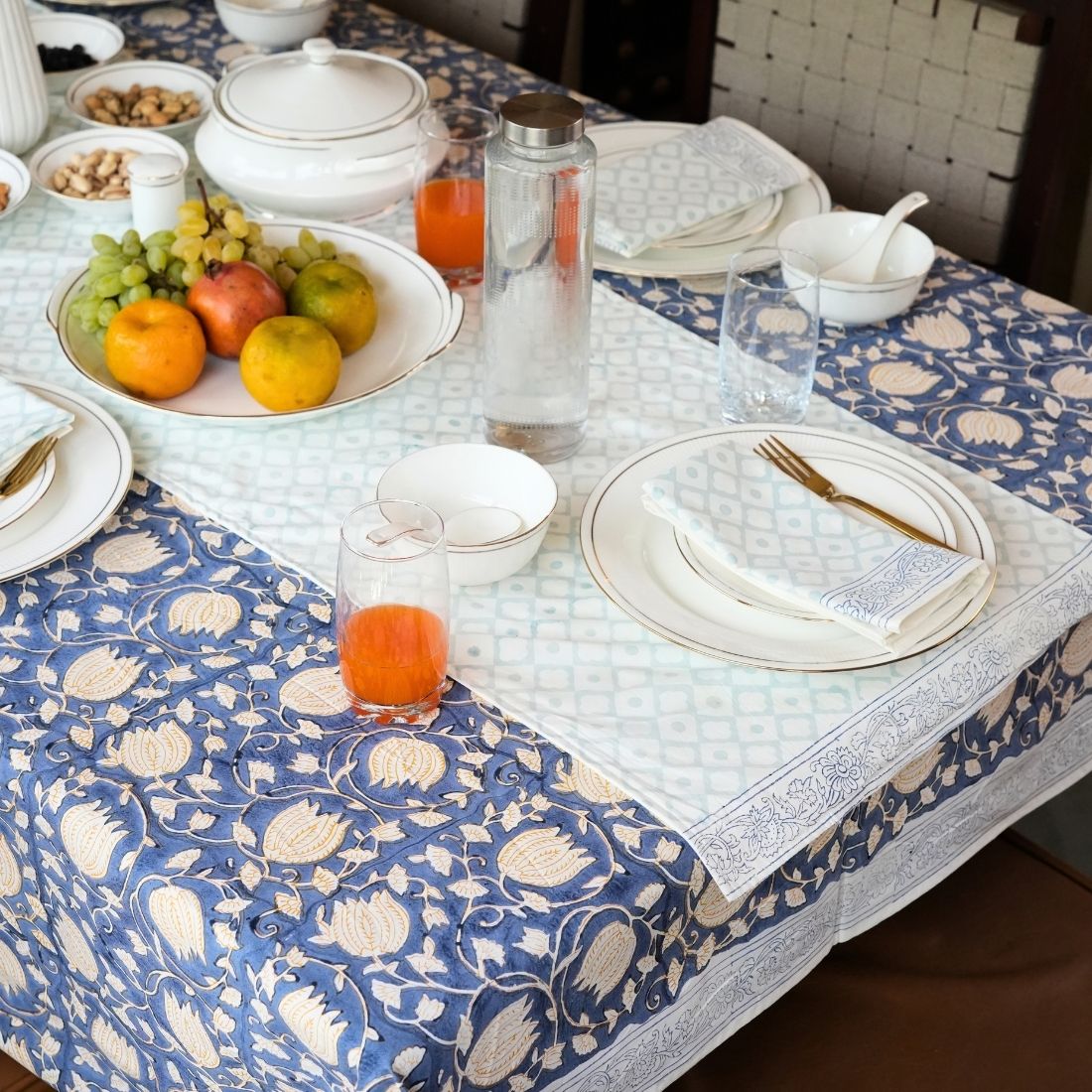 Dining table set with a blue and white patterned tablecloth, table runner, napkins, fruit plate, and glasses.