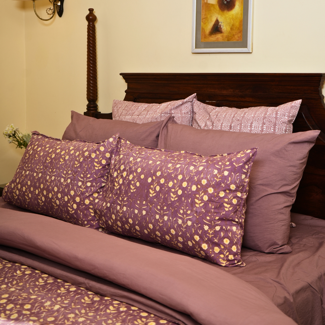 Bed with purple floral bedding and pillows against a beige wall.