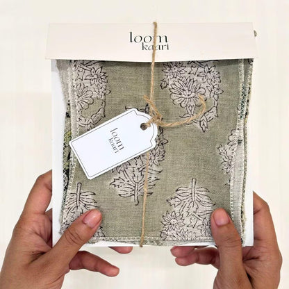 A person's hands holding a packaged textile product. The item appears to be a folded fabric piece or scarf in a sage green linen-like material with a beige botanical pattern, wrapped in clear plastic inside a white box with a kraft string and a white hangtag.