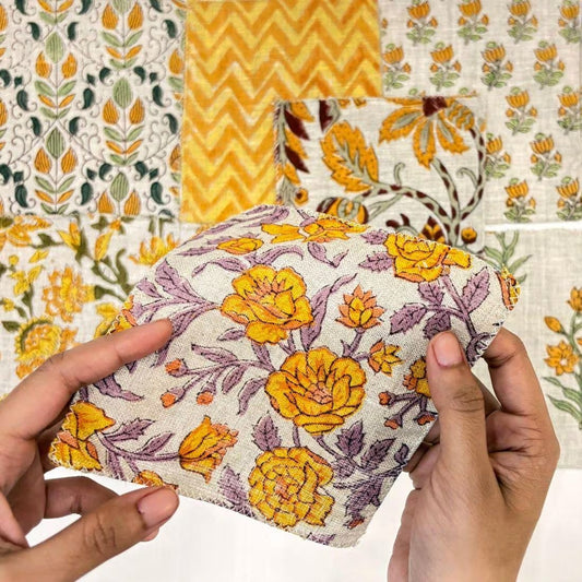 A person's hands holding a square fabric pouch with a yellow and purple floral print, set against a background of other patterned fabrics.