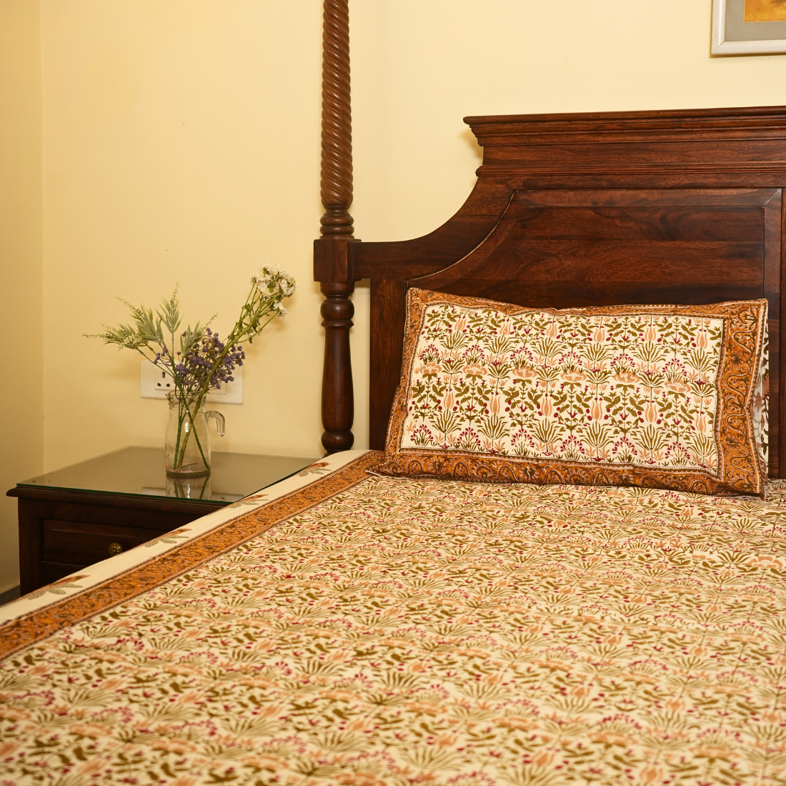 Bed with floral bedding and pillow, nightstand with flowers, and wooden headboard.