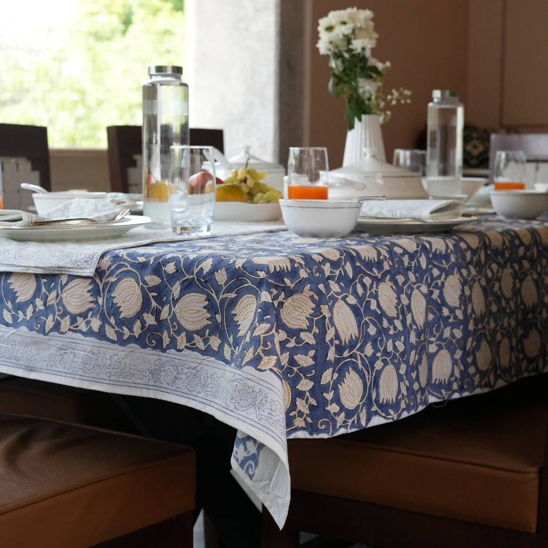 Dining table set with a blue floral table linen set, glasses, and fruit in a bright room.
