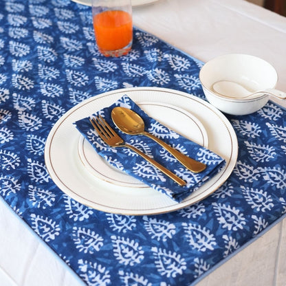 Table setting with blue and white patterned tablecloth, white plates, gold cutlery, and a blue napkin.