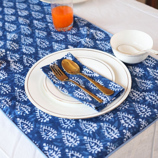 Table setting with blue and white patterned tablecloth, white plates, gold cutlery, and a blue napkin.