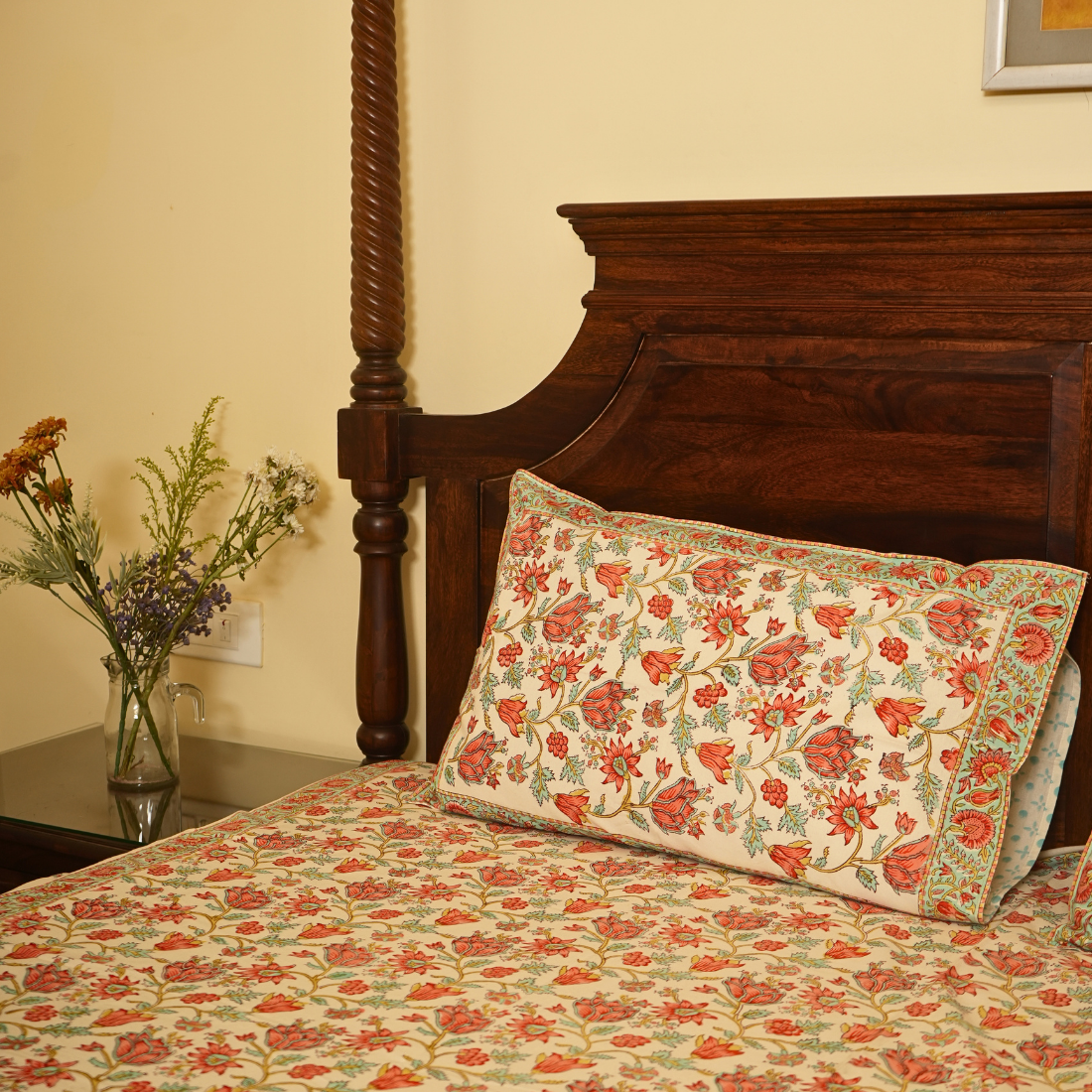 Floral-patterned bedspread and pillow on a wooden bed with a side table and flowers.