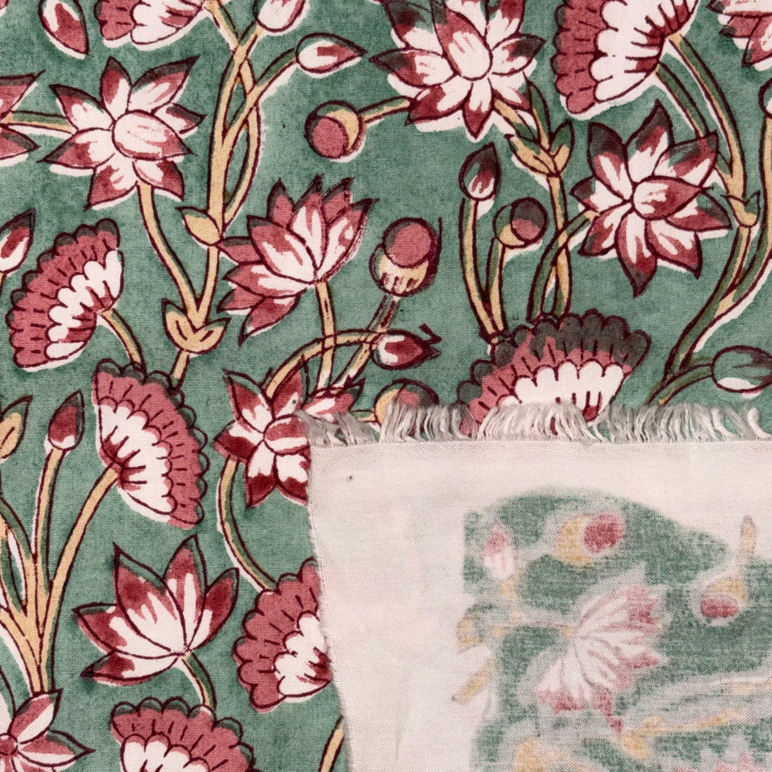 A close-up of a patterned textile or fabric with a sage green background, featuring pink and white lotus flowers with brown stems. A folded section at the bottom reveals a white backing with a matching printed floral design.