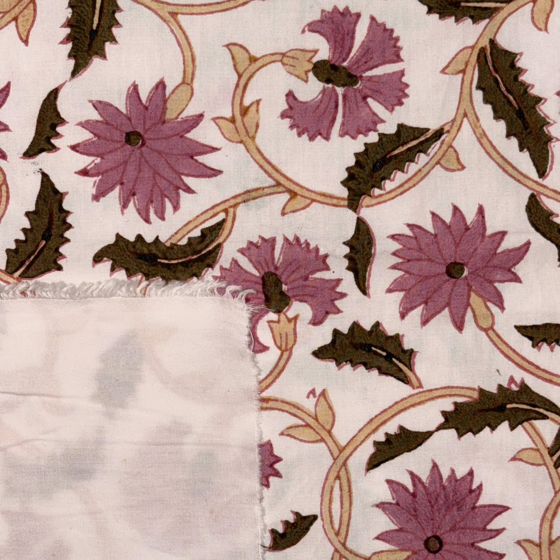 A close-up of a patterned textile, likely a tablecloth or napkin, featuring purple flowers and dark green leaves on a cream background with a folded over corner showing a light gray reverse side.