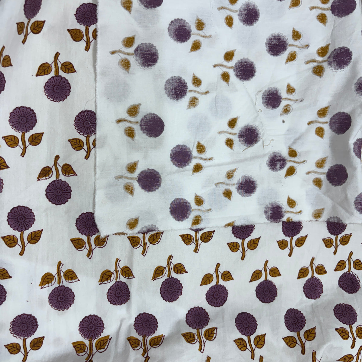 White fabric with a repeating block print pattern of purple flowers and brown leaves, showing both the printed side and the unprinted reverse side.