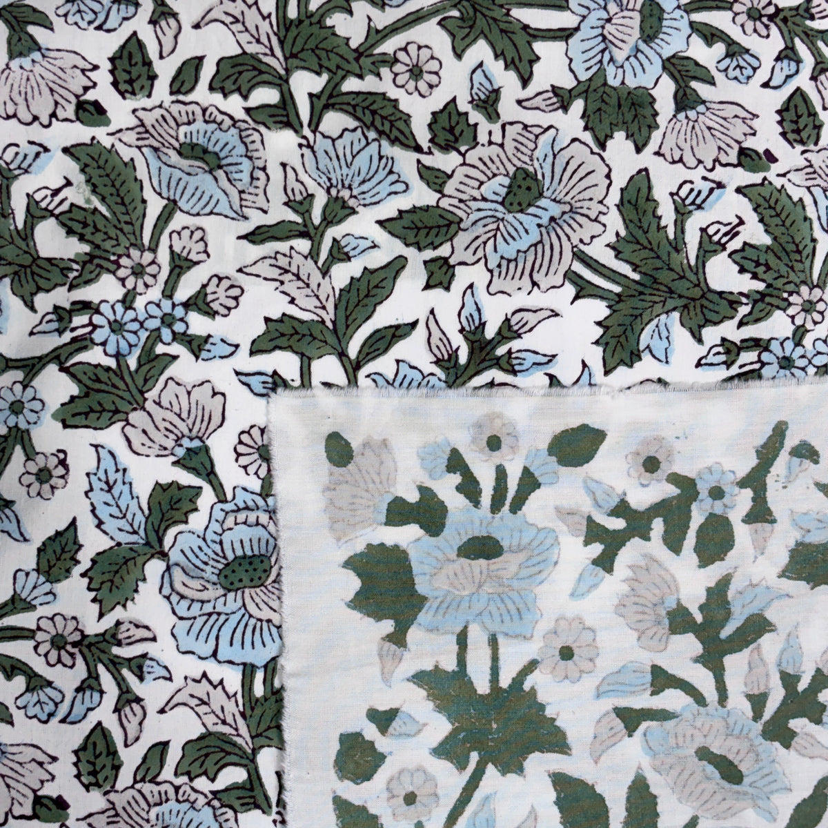 A close-up of a white fabric with a vintage floral block print pattern in muted green, light blue, and pale pink, with a folded corner showing the textured weave.