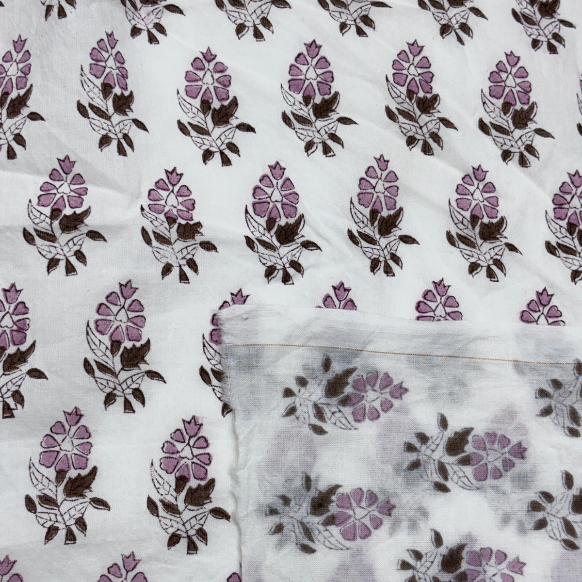 White fabric with a repeating hand-block print of purple and brown floral motifs, with a folded corner showing the texture and color variation.