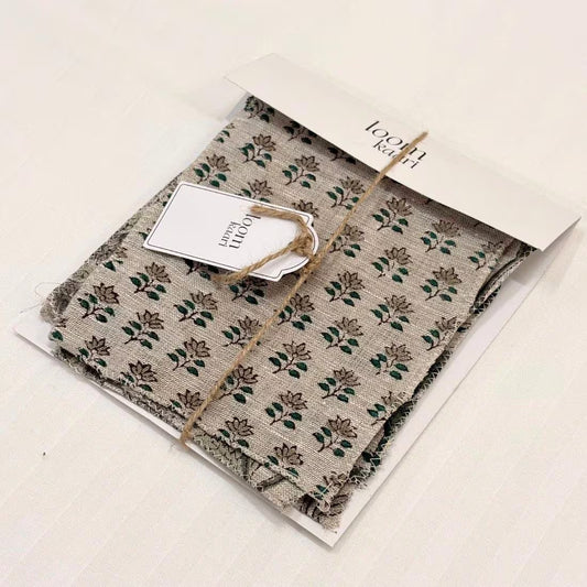 A folded patterned fabric item, likely a scarf or napkin, in a clear plastic sleeve with a white paper header card and a small white tag. The fabric has a beige or light gray base with a repeating green floral block-print style pattern.
