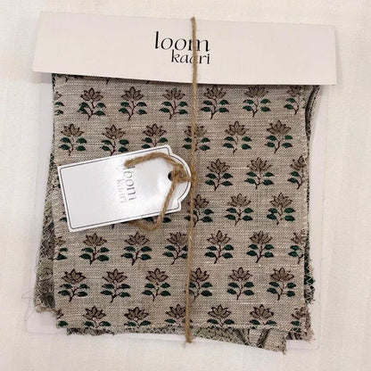 A packaged fabric sample with a beige base and dark green floral block print, wrapped in a kraft paper sleeve with a white paper header card reading 'loomkaari' and a small attached tag.
