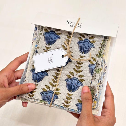 A pair of hands holding fabric swatches set bundle in its white packaging box. The fabric has a cream base with a blue floral tulip pattern and olive green leaves, tied with a jute string and a white paper tag.