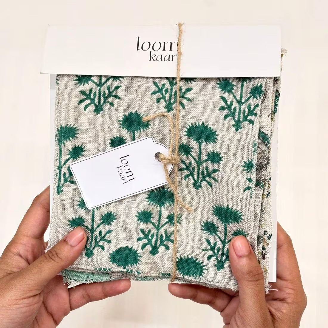 A pair of hands holding a packaged textile item with a cream base and green botanical print, wrapped in kraft paper with a white tag reading 'loom kareen' and 'handmade in India'.