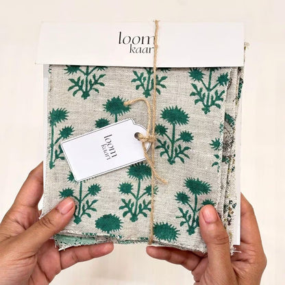 A pair of hands holding a packaged textile item with a cream base and green botanical print, wrapped in kraft paper with a white tag reading 'loom kareen' and 'handmade in India'.