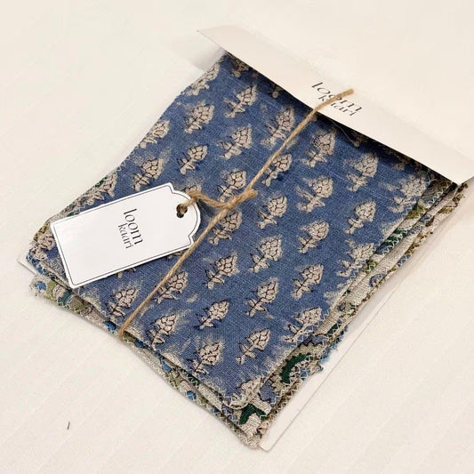 A folded piece of blue fabric with a repeating beige botanical block print, tied with twine and attached to a white paper tag reading 'loomi fabric,' placed on a white background.