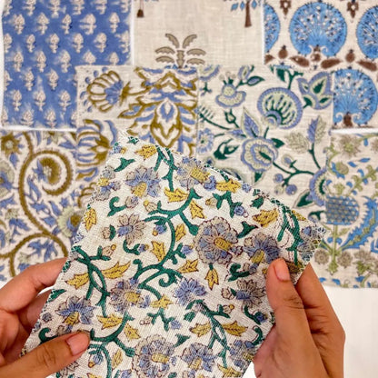 A close-up of hands holding a square piece of patterned fabric in front of a larger matching fabric swatch display. The fabric features a floral and vine print in blue, purple, green, and yellow on an off-white background.