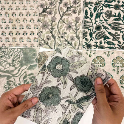 A close-up of hands holding a patterned fabric sample, with four other matching patterned samples arranged around it. The designs feature muted green botanical and floral motifs on an off-white background.