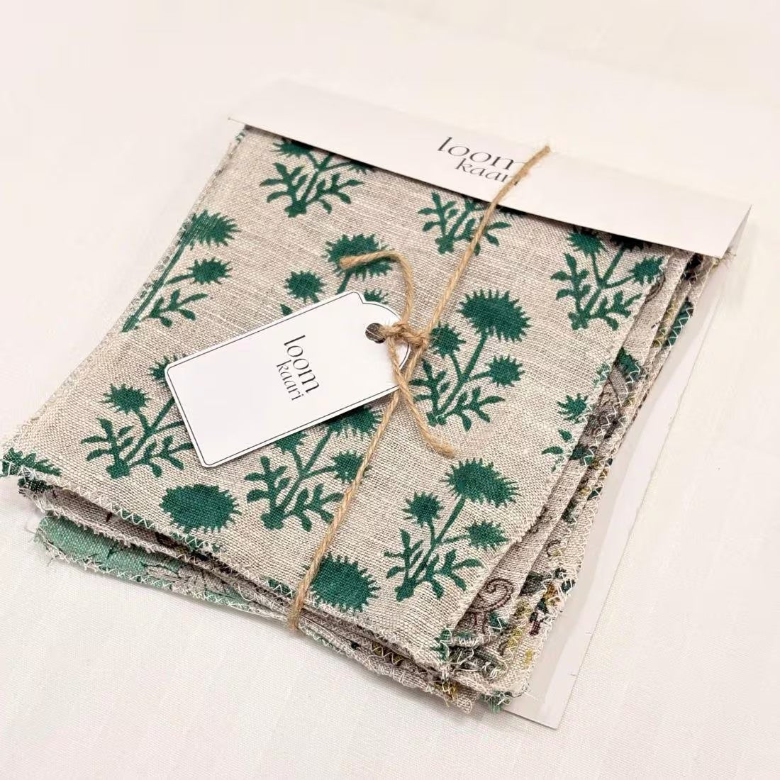 A stack of linen fabric squares with a green floral thistle pattern on a beige linen background, tied with twine and a paper tag. The brand name 'loom knit' is visible on the tag and the paper header.