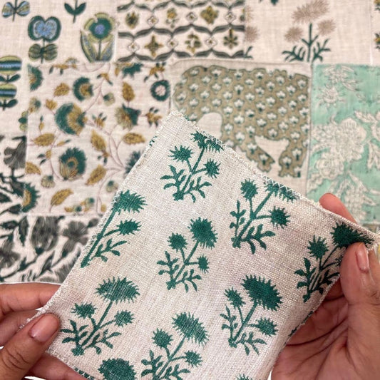 A close-up of hands holding a square piece of cream-colored fabric with a green botanical thistle pattern, set against a background of a larger patchwork quilt made of various floral and geometric fabrics.