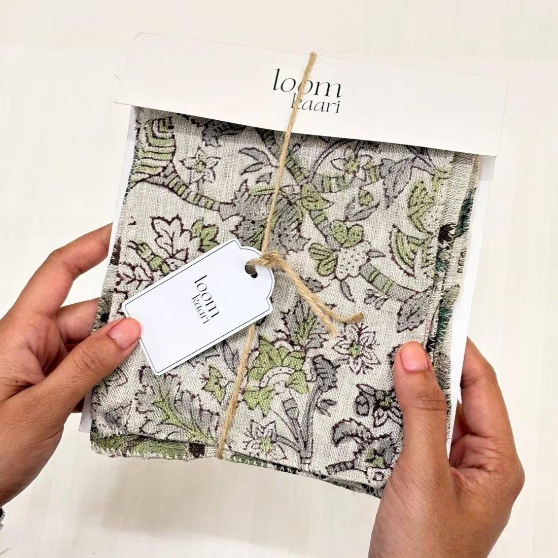 A person's hands holding a packaged fabric item with a botanical pattern. The item is wrapped in a kraft paper sleeve with a white tag that reads 'loomkaari'
